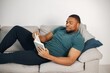 Black guy lay on a couch in living room with a book
