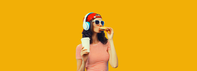 Wall Mural - Stylish young woman listening to music in headphones eating burger fast food on yellow background