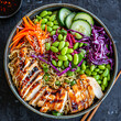 An aerial view of a colorful Asian noodle salad arranged in a large bowl, with cooked noodles, shredded carrots, cucumber slices, red cabbage, edamame beans, and sliced grilled chicken, tossed in a se