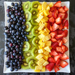 An aerial shot of a fresh fruit salad arranged in a geometric pattern, showcasing a colorful assortment of sliced strawberries, blueberries, kiwi, pineapple, and grapes on a white serving platter.
