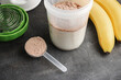 Whey protein powder with a chocolate flavor in a plastic measuring spoon, shaker with a milk and bananas on a dark background, process of making protein shake drink