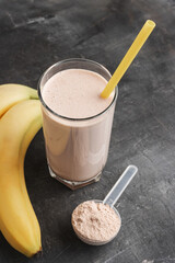 Wall Mural - Blended chocolate protein drink in a glass with yellow straw, plastic measuring spoon with protein powder and bananas on a dark background