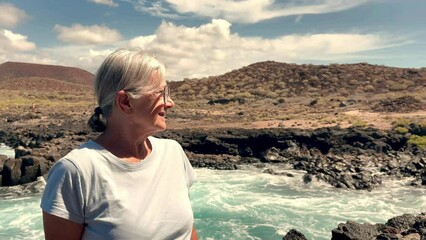 Sticker - Smiling senior white-haired woman sitting on a rocky beach looking the waves crashing on the rocks. Elderly female in retirement or vacation enjoying a sunny day and freedom outdoors