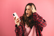 Photo portrait of panicking brunette woman with open mouth touching head holding phone in one hand isolated on pink colored background. OMG!