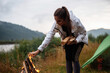 Positive young woman preparing bonfire. Joyful woman enjoying of the camping at the nature near her tent. Girl alone at the forest