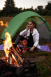 Camping at the forest. Happy backpacker woman sitting in entrance of tourist tent at campfire near forest under sky, enjoying beautiful views