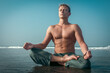 A shirtless young man sits in a meditative lotus pose on the wet sand of a beach, with the ocean waves crashing behind him.