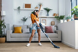 Fototapeta Lawenda - Shot of young happy woman listening and dancing to music while cleaning the living room floor with a vaccum cleaner