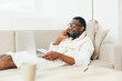 Smiling African American Freelancer Working on Laptop on Sofa in Home Office, Talking on Phone in Bathrobe