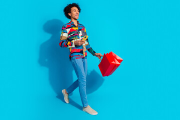 Sticker - Full size photo of handsome young guy hold telephone walking shopping bags dressed stylish print garment isolated on blue color background