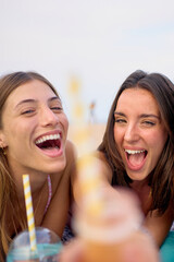 Wall Mural - Vertical. Group of attractive Caucasian gen z women looking excited at camera summer day. They holding unfocused natural smoothies foreground. Nice female friends enjoy vacation posing lying outdoors