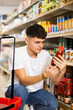 young man meticulously chooses tomato sauce in supermarket.