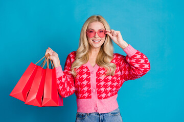 Poster - Portrait of adorable person dressed knitwear jumper touch pink sunglass holding new outfit in bags isolated on blue color background