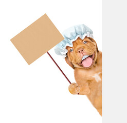 Wall Mural - Happy Mastiff puppy with funny big teeth wearing shower cap looking from behind empty white banner and showing empty placard. isolated on white background