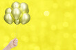 Female hand holding bunch of shiny golden or yellow balloons on blurred yellow background. Empty space for text