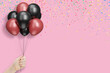 Female hand holds bunch of red and black balloons on pink background with confetti. Empty space for text