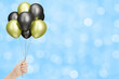 Female hand holds bunch of golden and black balloons on blurred blue background. Empty space for text