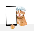 Mastiff puppy wearing shower cap with cream on it face holds big smartphone with white blank screen in it paw above empty white banner. isolated on white background