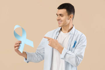 Wall Mural - Young man pointing at blue ribbon on beige background. Prostate cancer awareness concept