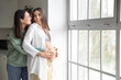 © Pixel-Shot - Young lesbian woman hugging her pregnant wife near window in kitchen