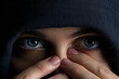 Close-up of a person's eyes peering out from under a dark hood. Model holding hands and fingers near nose