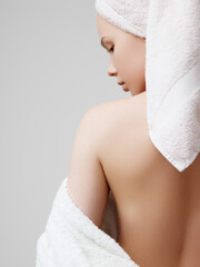 Back view of pretty female with towel on head and in bathrobe posing, looking over shoulder. Portrait of woman enjoying time after fresh shower, isolated on grey background. Beauty, skincare concept.