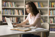 © fizkes - Focused young girl student prepare for exam in university library, use laptop, browse important information on internet, send email, work on essay, write coursework. E-learning, education, modern tech