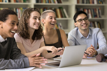 Wall Mural - Happy generation Z students sit at desk in college library, look away, engaged in teamwork, exams preparation, distracted from study, laughing, joking, involved in communication and knowledge getting