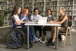 Pretty student girl with disability sits in wheelchair talking to interracial schoolmates, studying together, joking, laughing, make joint project, engaged in teamwork having good friendly relations