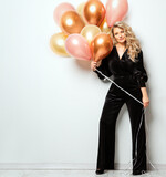 Fototapeta  - Beautiful Woman with Balloons over White background. Birthday Party Time. Fashion Model with Curly Hairstyle in Black Suit