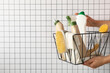 Basket with bottles of detergents and brushes in hands on light background, space for text