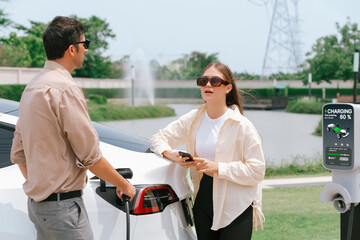 young couple recharge electric car's battery from charging station in outdoor green city park in spr