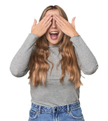 Wall Mural - Studio portrait of a blonde Caucasian woman covers eyes with hands, smiles broadly waiting for a surprise.