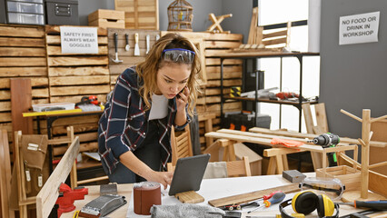 Canvas Print - Portrait of a beautiful young hispanic woman carpenter, engrossed in online business on her touchpad while casually talking on smartphone amidst the rustic charm of her carpentry workshop