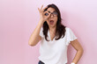 Middle age hispanic woman wearing casual white t shirt and glasses doing ok gesture shocked with surprised face, eye looking through fingers. unbelieving expression.