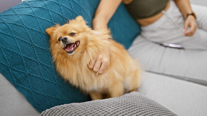 Sticker - Beautiful woman sitting together with her pet dog on a sofa at home, indoor portrait capturing the touching moment of her hands holding its paw in their cozy living room interior.