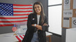 © Krakenimages.com - A confident hispanic woman stands with crossed arms in an american electoral college room with a flag and voting signs.