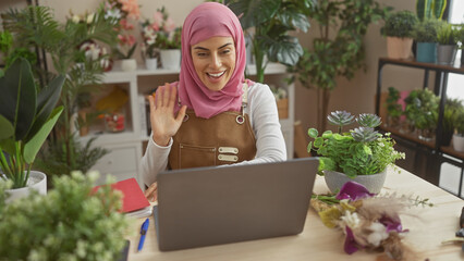 Wall Mural - Smiling woman in hijab waving during video call at home office surrounded by plants