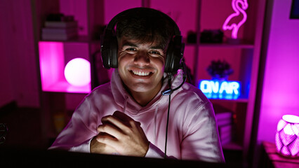 Canvas Print - Smiling young hispanic man streamer sporting headphones, blissfully immersed in the pulse of a night time gaming stream at his indoors gaming room