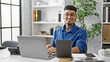 © Krakenimages.com - Confident young latin man, smiling happily while working online using his laptop and touchpad at a professional business office