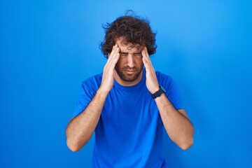 Poster - Hispanic young man standing over blue background with hand on head, headache because stress. suffering migraine.