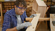 © Krakenimages.com - Serious young hispanic carpenter in glasses, fashioning a masterpiece, touching and feeling his plank's texture in a cozy indoor carpentry workshop.