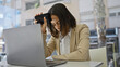 © Krakenimages.com - A stressed young hispanic woman at a desk with a laptop holding her phone to her temple in a beige office environment.