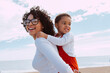 Smiling beautiful mother and her daugther enjoy together piggyback on beach, Love, vacation, happy family, ocean and tourism concept. Child hugs woman outdoor, mockup space.