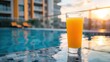 a glass of orange juice placed on the pool's edge, with a luxurious hotel and shimmering swimming pool in the blurred background, set against the backdrop of a sunny day.