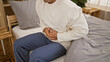 © Krakenimages.com - Young hispanic man, a handsome guy, caught in the agony of a stomach ache, sitting on bed, clutching his abdomen. his portrait in a painful moment, suffering in the heart of his apartment bedroom.