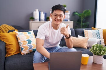 Wall Mural - Young asian man using laptop at home sitting on the sofa doing happy thumbs up gesture with hand. approving expression looking at the camera showing success.