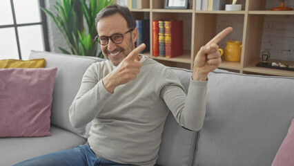 Wall Mural - Smiling bearded middle-aged man pointing sideways while sitting on a couch in a modern living room.