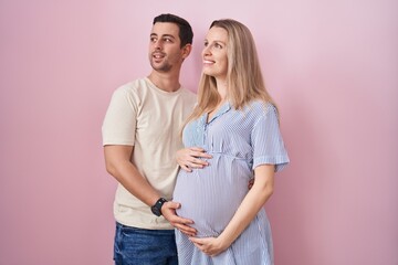 Young couple expecting a baby standing over pink background looking away to side with smile on face, natural expression. laughing confident.