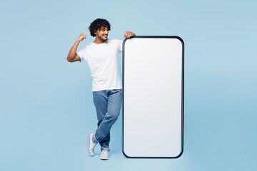 Wall Mural - Full body young Indian man wear white t-shirt casual clothes big huge blank screen mobile cell phone smartphone with area do winner gesture isolated on plain pastel blue background. Lifestyle concept.